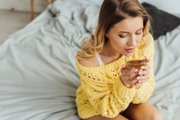 Schöne junge Frau mit geschlossenen Augen sitzt auf dem Bett und hält eine Tasse Tee — Stockfoto