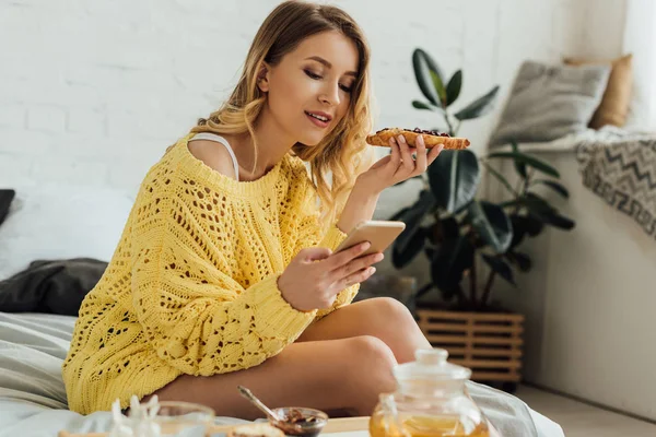 Hermosa chica en suéter de punto celebración de tostadas y el uso de teléfono inteligente durante el desayuno en casa - foto de stock