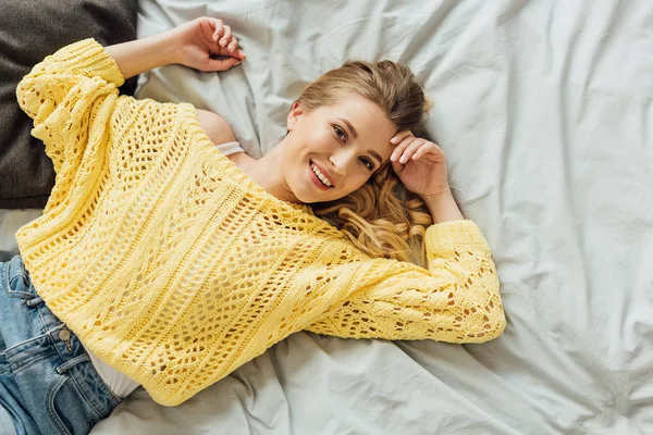 Draufsicht der schönen jungen Frau im Strickpullover, die auf dem Bett liegt und in die Kamera schaut — Stockfoto