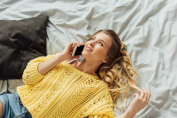 Hermosa chica sonriente acostada en la cama y hablando en el teléfono inteligente - foto de stock