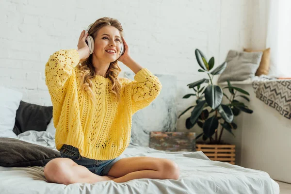 Schöne, glückliche junge Frau mit Kopfhörern, die auf dem Bett sitzt und zu Hause Musik hört — Stockfoto