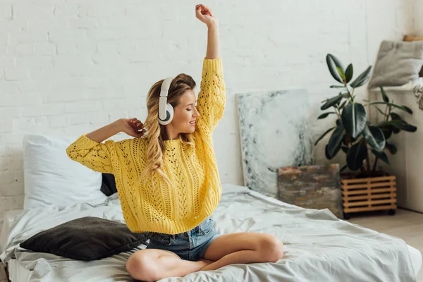 Bella giovane donna sorridente in cuffia seduta sul letto, che si allunga e ascolta musica a casa — Foto stock