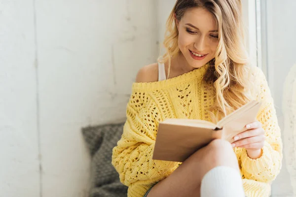 Bela sorridente jovem mulher ler livro enquanto sentado na soleira da janela com espaço de cópia — Fotografia de Stock