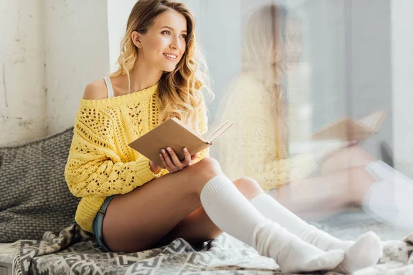 Bela sorridente jovem segurando livro enquanto sentado na soleira da janela com espaço de cópia — Fotografia de Stock