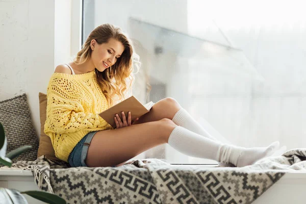 Красивая улыбающаяся молодая женщина читает книгу, сидя на подоконнике с копировальным пространством — стоковое фото