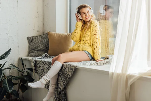 Красивая улыбающаяся молодая женщина в наушниках сидит на подоконнике и слушает музыку дома — стоковое фото