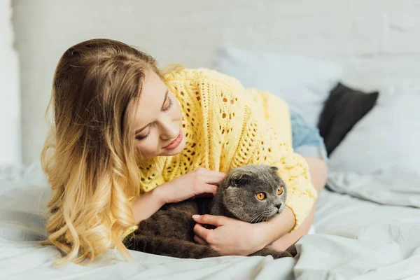 Schönes Mädchen im Strickpullover liegt im Bett und umarmt schottische Faltkatze — Stockfoto