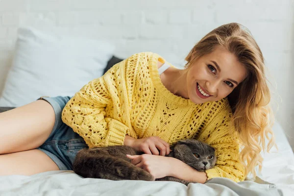 Bela menina sorridente em camisola de malha olhando para a câmera e abraçando escocês dobra gato na cama — Fotografia de Stock