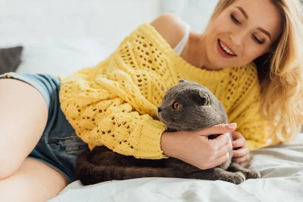 Foco seletivo de bela menina sorridente em suéter de malha deitado na cama e abraçando escocês gato dobra — Fotografia de Stock