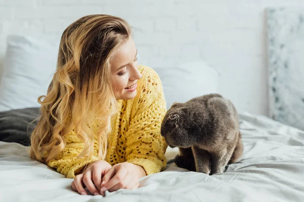 Hermosa chica rubia sonriente en suéter de punto acostado en la cama con gato pliegue escocés en casa - foto de stock