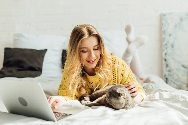 Schönes lächelndes Mädchen, das mit schottischer Faltkatze im Bett liegt und zu Hause Laptop benutzt — Stockfoto
