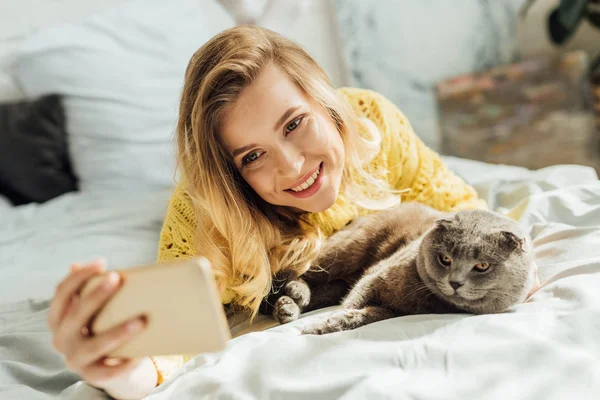 Selective focus of beautiful smiling young woman taking selfie on smartphone while lying in bed with scottish fold cat — Stock Photo