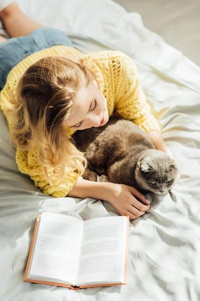 Vue grand angle de belle jeune femme couchée au lit avec chat pliant écossais et livre — Photo de stock