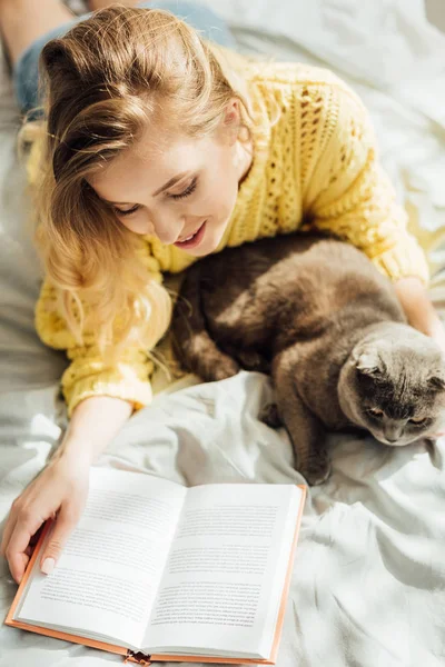 Visão de alto ângulo de belo sorriso jovem leitura livro enquanto deitado na cama com escocês dobra gato — Fotografia de Stock