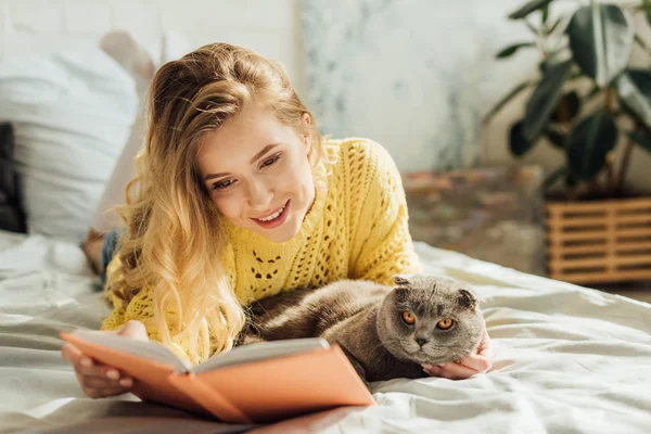 Красивая улыбающаяся молодая женщина читает книгу, лежа в постели со шотландским складным котом — стоковое фото