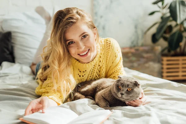 Красивая молодая женщина, глядя в камеру и держа в руках книгу, лежа в постели с кошкой со скоттишем — стоковое фото