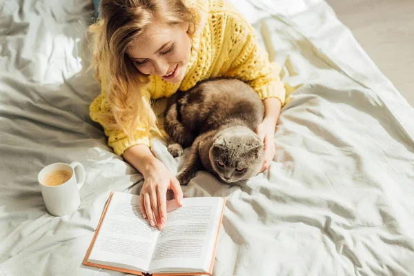 Hochwinkelaufnahme der schönen jungen Frau, die Buch liest, während sie mit schottischer Faltkatze im Bett liegt — Stockfoto