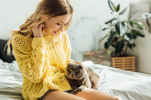 Selective focus of beautiful smiling blonde young woman sitting on bed with scottish fold cat — Stock Photo