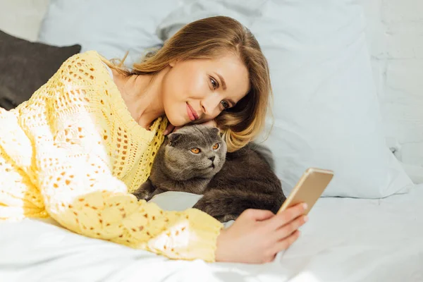 Hermosa joven mujer tomando selfie en smartphone mientras acostado en cama con escocés plegable gato - foto de stock