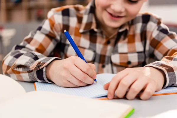 Vista parcial del alumno sonriente con camisa a cuadros escribiendo en cuaderno durante la lección - foto de stock