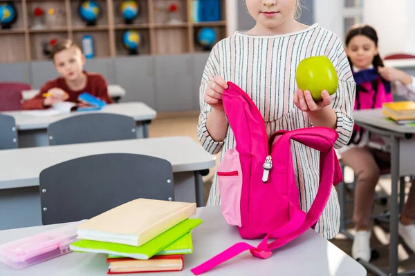 Vista recortada de colegiala sosteniendo manzana verde y mochila rosa en el aula — Stock Photo