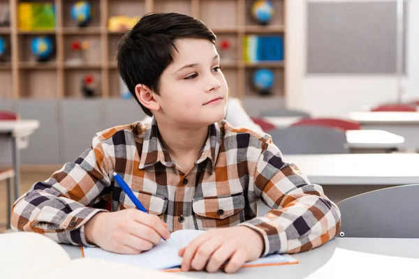 Pensive brunette pupil in checkered shirt writing in notebook during lesson — Stock Photo