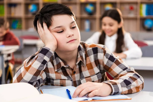 Upset schoolboy with notebook sitting at desk in classroom — Stock Photo