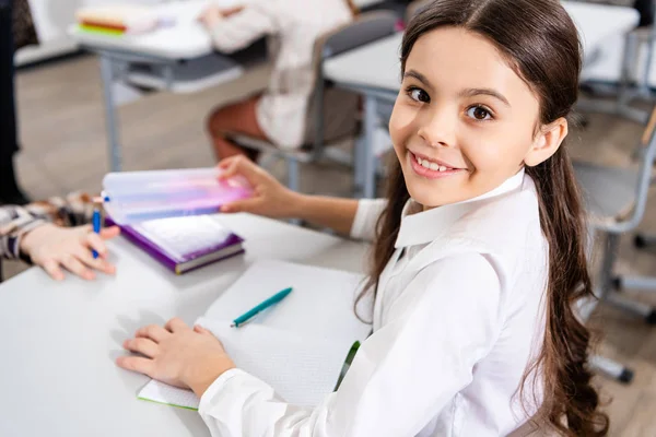 Alegre colegiala sonriente sentada en el escritorio y mirando a la cámara en el aula - foto de stock