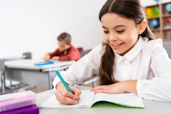 Lächelndes Schulmädchen schreibt während des Unterrichts im Klassenzimmer in Notizbuch — Stockfoto