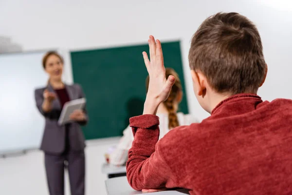 Back view of schoolboy raising hand during lesson in classroom — Stock Photo