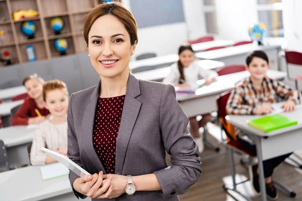 Smiling teacher with digital tablet standing in front of pupils and looking at camera — Stock Photo