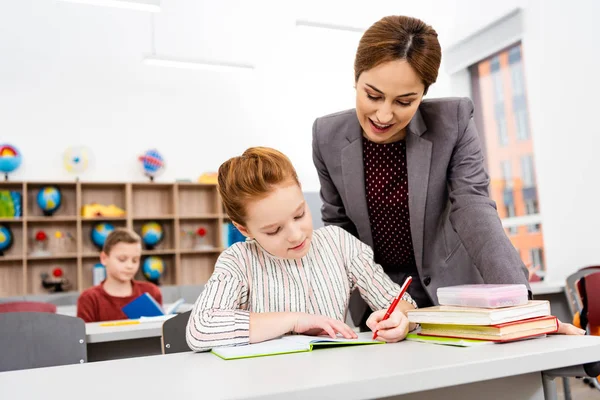 Teacher standing near desk and explaining lesson to pupil in classroom — Stock Photo