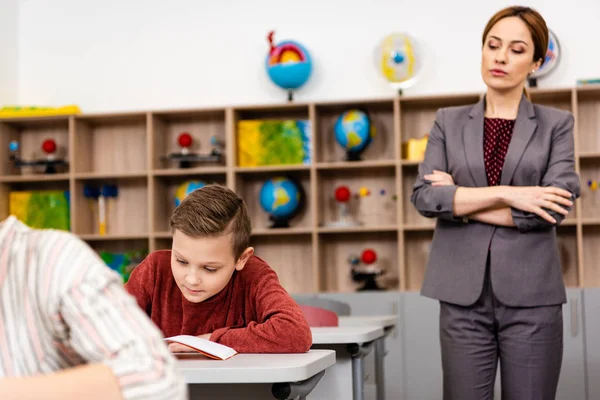 Serious teacher in suit standing with crossed arms and looking at pupils — Stock Photo