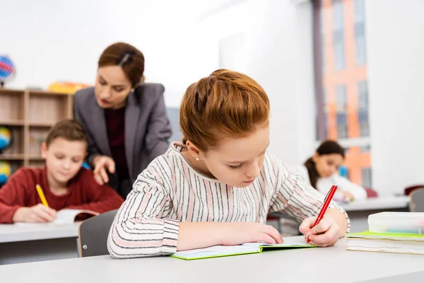 Linda colegiala de jengibre escribiendo en cuaderno durante la lección en el aula - foto de stock