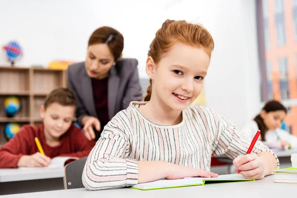 Cute ginger schoolgirl writing in notebook during lesson in classroom — Stock Photo