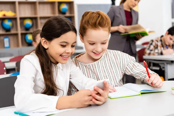 Smiling schoolgirls sitting at desk and using smartphone during lesson in classroom — Stock Photo