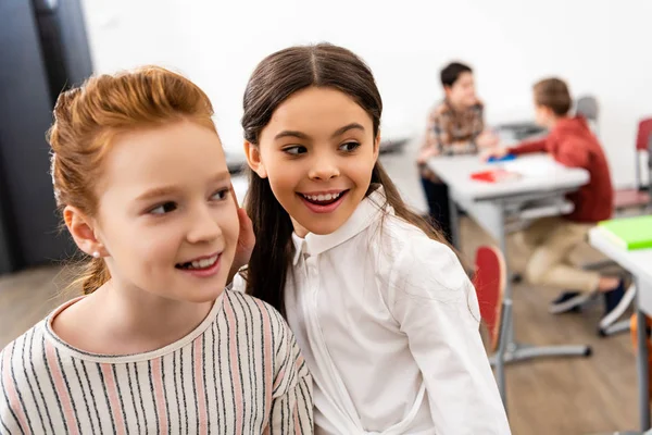 Dos colegialas lindas sonriendo y mirando hacia otro lado en el aula durante el freno - foto de stock