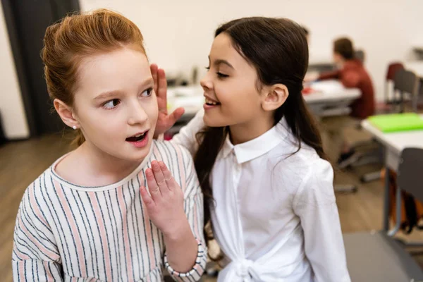 Two cute schoolgirls smiling and sharing gossips in classroom during brake — Stock Photo
