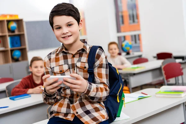 Elève souriante en chemise à carreaux avec sac à dos en utilisant un smartphone en classe — Photo de stock