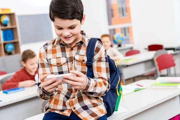 Smiling pupil in checkered shirt with backpack using smartphone in classroom — Stock Photo