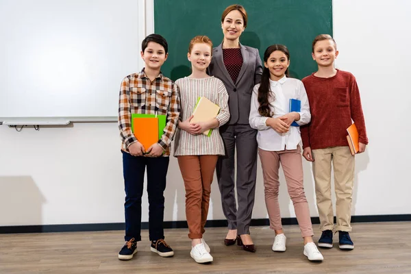 Professora e alunos com cadernos em pé em frente ao quadro negro — Fotografia de Stock