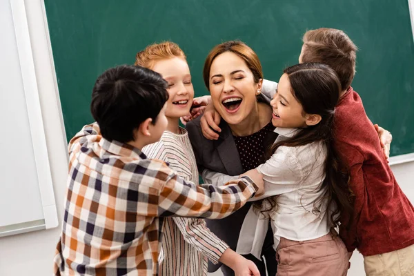 Allievi felici abbracciare insegnante davanti alla lavagna in classe — Foto stock