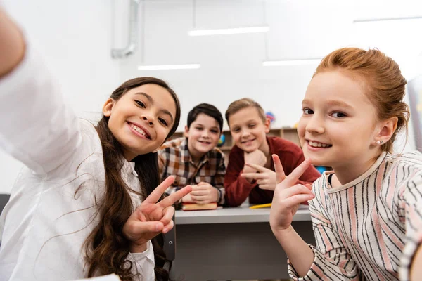 Alegre colegiala tomando selfie con amigos y mostrando signo de paz en el aula - foto de stock