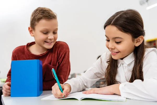 Smiling schoolgirl talking with friend while writing in notebook in classroom — Stock Photo