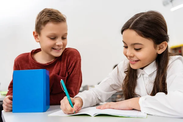 Studentessa sorridente che parla con un amico mentre scrive nel taccuino in classe — Foto stock