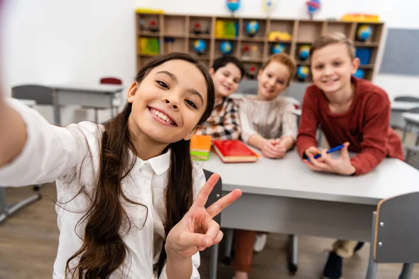 Écolière joyeuse prenant selfie avec des amis et montrant signe de paix en classe — Photo de stock