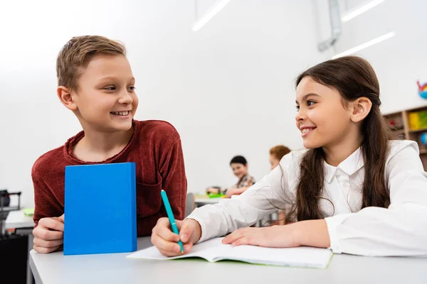 Smiling schoolgirl talking with friend while writing in notebook in classroom — Stock Photo