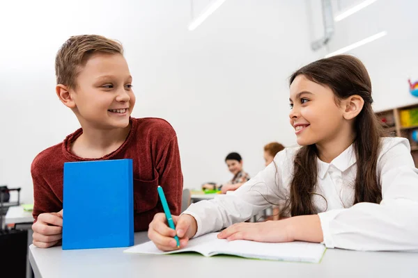 Estudante sorrindo conversando com o amigo enquanto escrevia no caderno em sala de aula — Fotografia de Stock