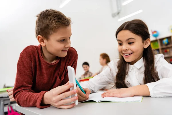 Escolar sonriente mostrando smartphone a amigo en el escritorio en el aula - foto de stock