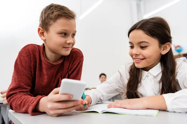 Escolar sonriente mostrando smartphone a amigo en el escritorio en el aula - foto de stock